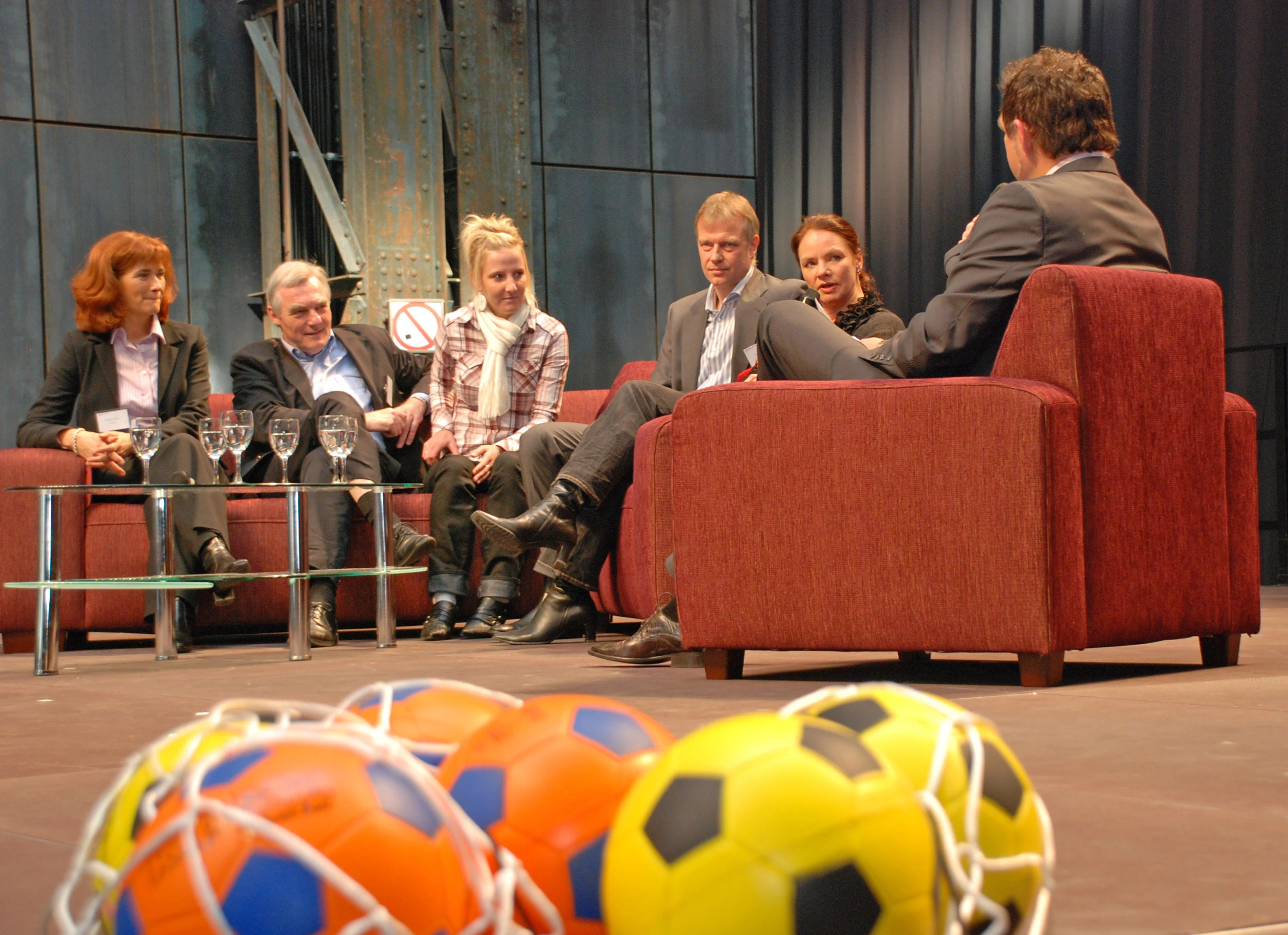 Gästerunde auf roter Couch mit Fußbällen im Vordergrund.