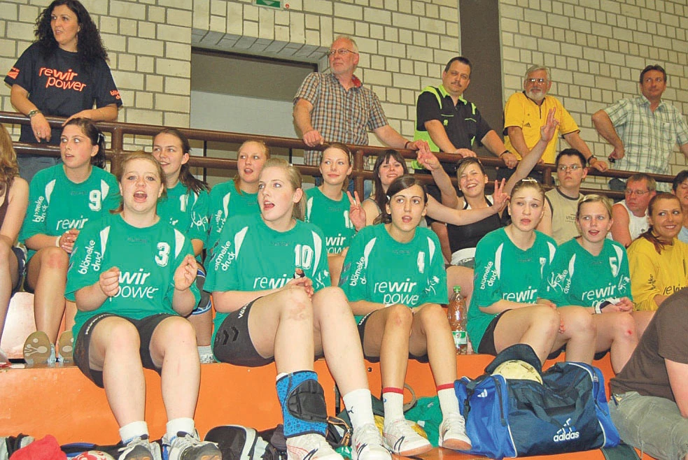 Applaudierende Handballerinnen in grünen Trikots auf der Tribüne sitzend