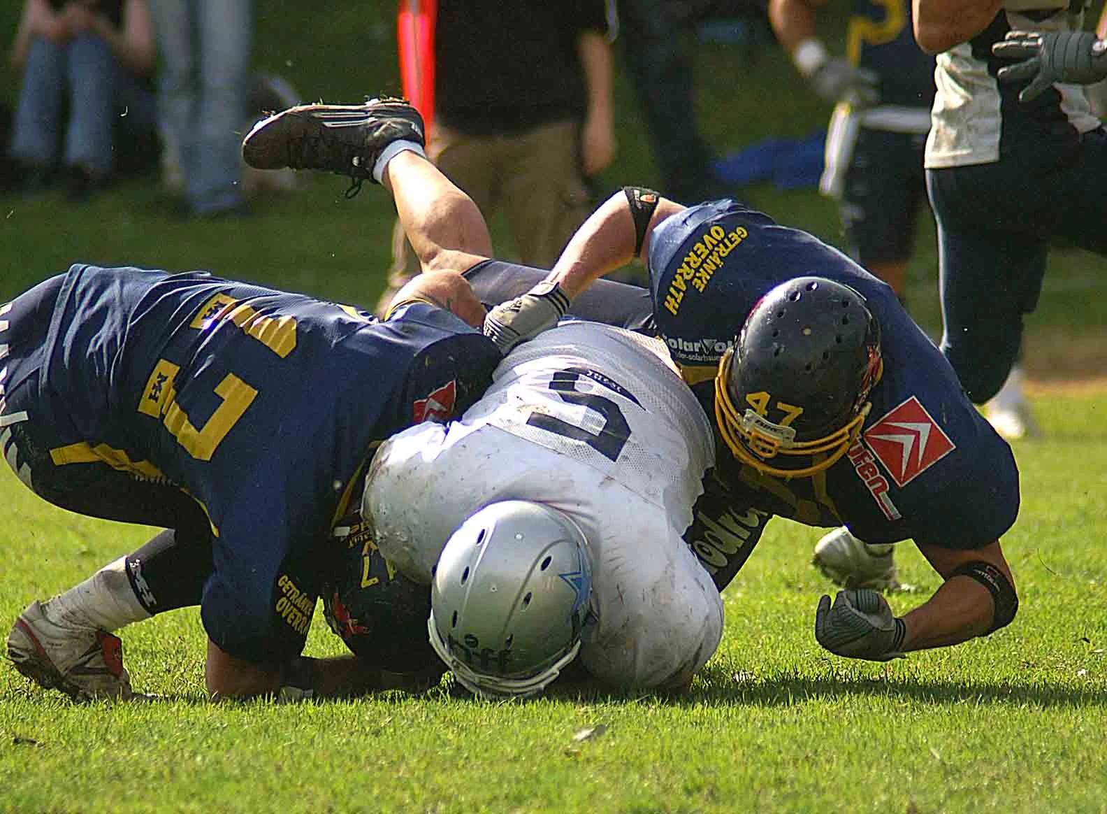 Drei American Footballer kämpfen auf der Wiese liegend um das Ei.