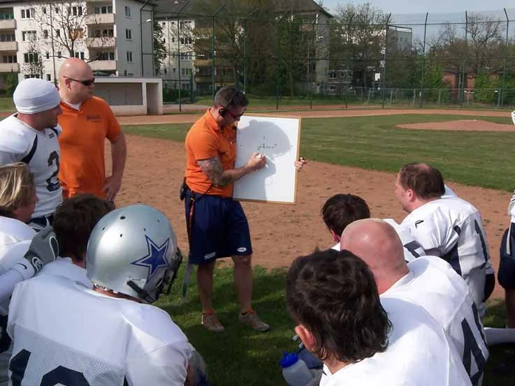 Trainer steht mit Headset und Schautafel vor seinen Spielern