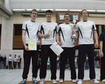 Vier Männer in schwarzen Hosen und weißen Triktos mit Pokal in den Händen auf dem Siegerpodest