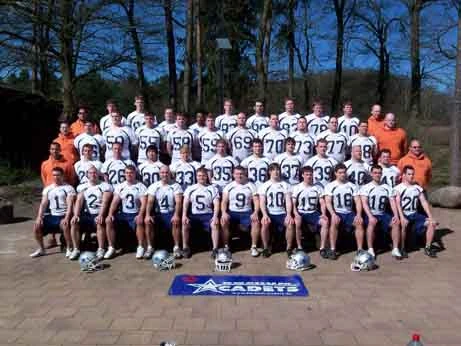 Die American Footballer haben sich in vier Reihen mit ihren Trainern aufgestellt und präsentieren sich in weißen Trikots und blauen Hosen