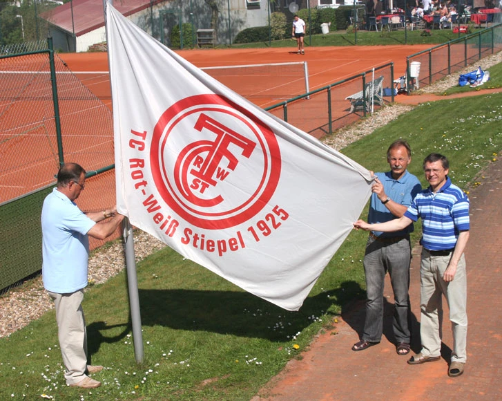 Drei Männer halten die weiße Fahne mit dem roten Vereinslogo des Stiepeler Tennisclubs. Im Hintergrund liegen die Tennisplätze