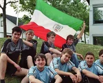 Vier junge Prellballer in schwarzen Trikots mit grün-weiß-roter Vereinfarbe. Vor ihnen vier junge Prellballer in hellblauen Trikots. Alle sitzen auf einer Wiese.